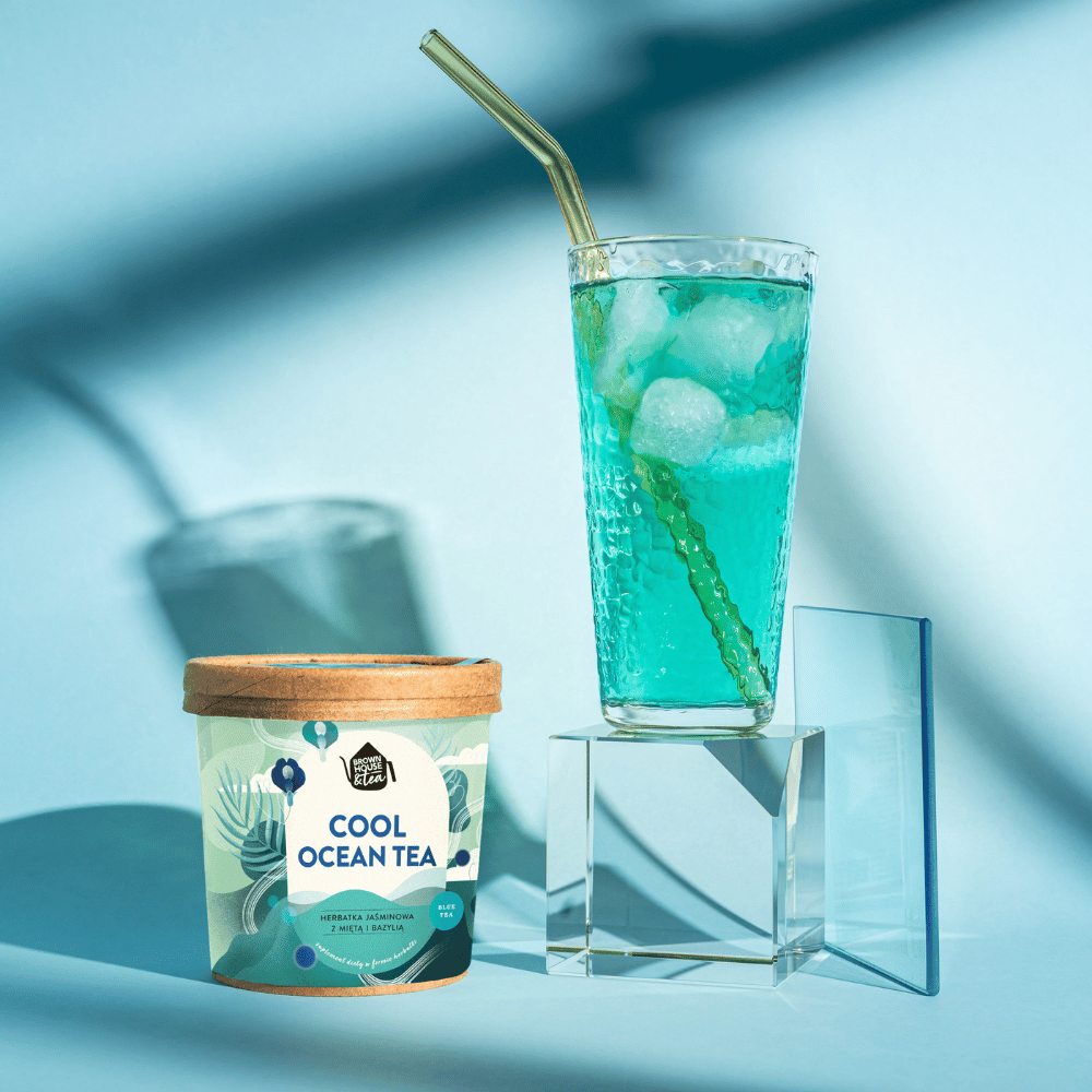 herbata cool ocean iced tea w pojemniku za nią na przezroczystym podeście zaparzona na zimno niebieska herbata z kostkami lodu i słomką w wysokiej szklance. całość na jasnoniebieskim tle z lekkim cieniem.