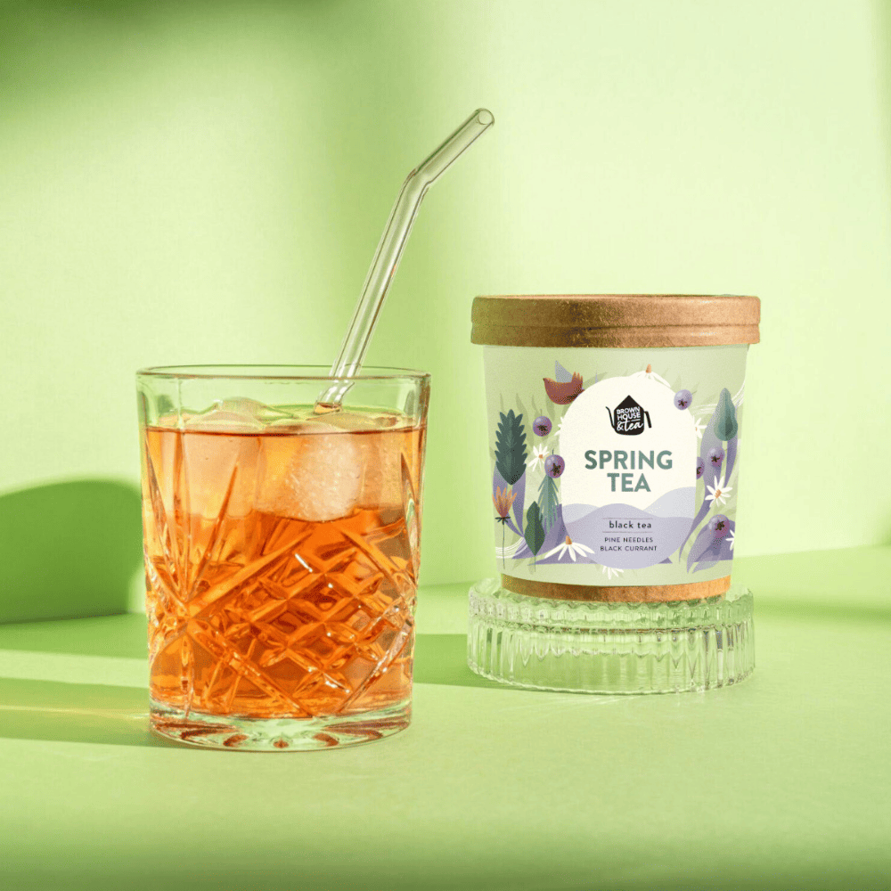 herbata spring tea w pojemniku obok w szklance zaparzona herbata na zimno, szklanka kryształ w której widać kostki lodu, zanurzona przezroczysta słomka, wszystko na jasnozielonym tle.