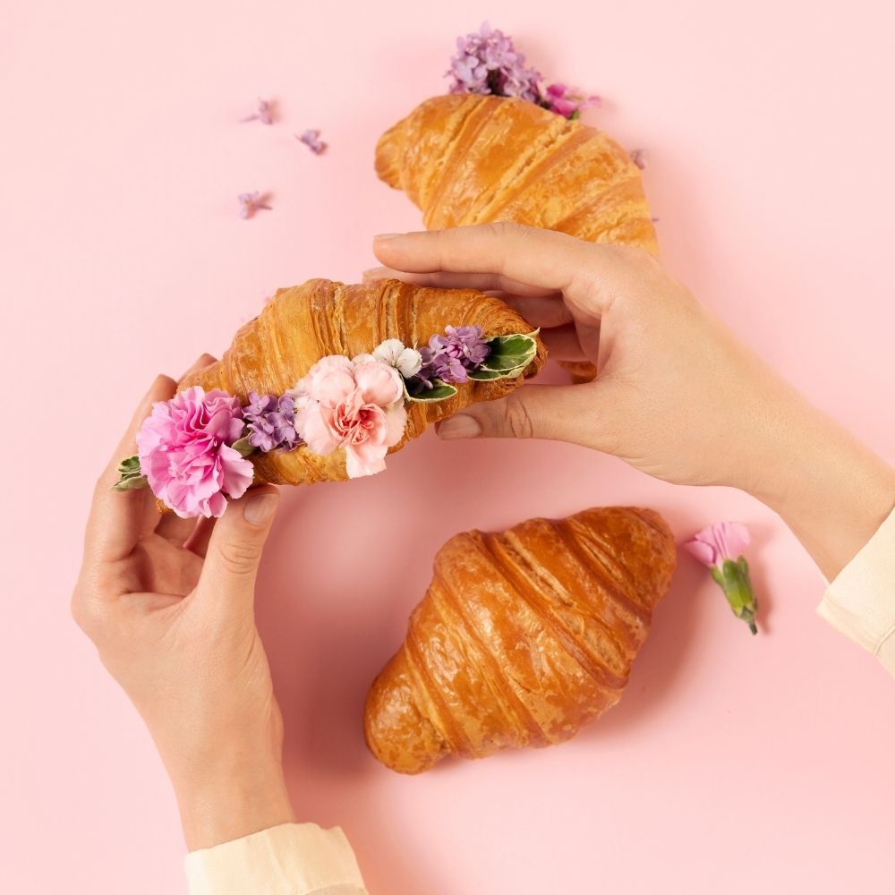 rogaliki francuskie croissant z kwiatami na jasnoróżowym tle, jeden rogalik trzymany w dłoniach